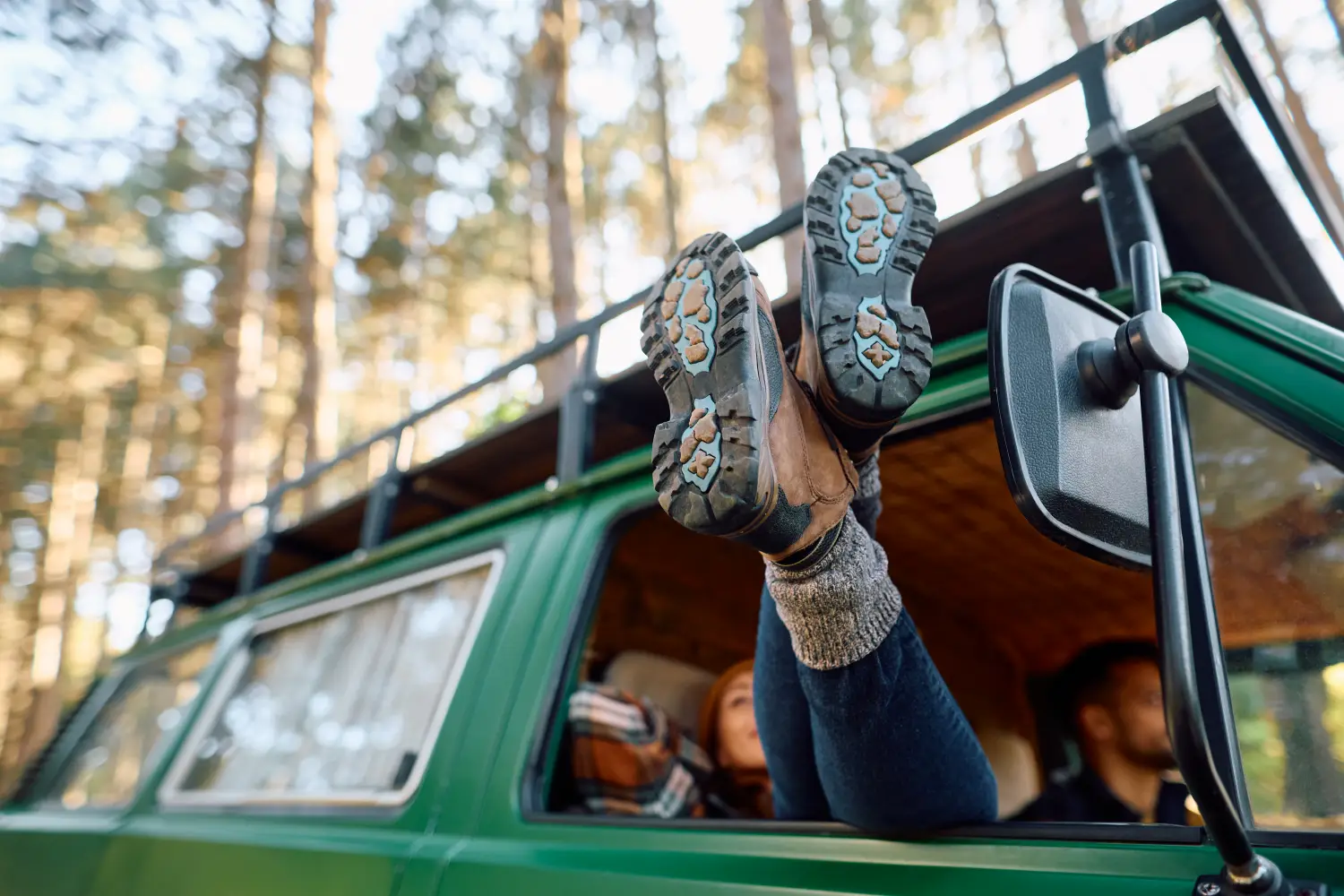 O persoană își întinde picioarele încălțate în ghete de drumeție pe fereastra deschisă a unei autoutilitare de camping de culoare verde, în timp ce se relaxează în pădure.